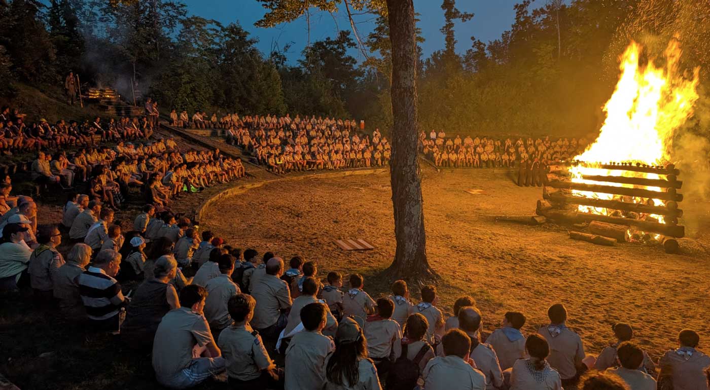 A large bonfire rages, surrounded by a crowd of OA members sitting on levels of elevated ground which mimic an amphitheater