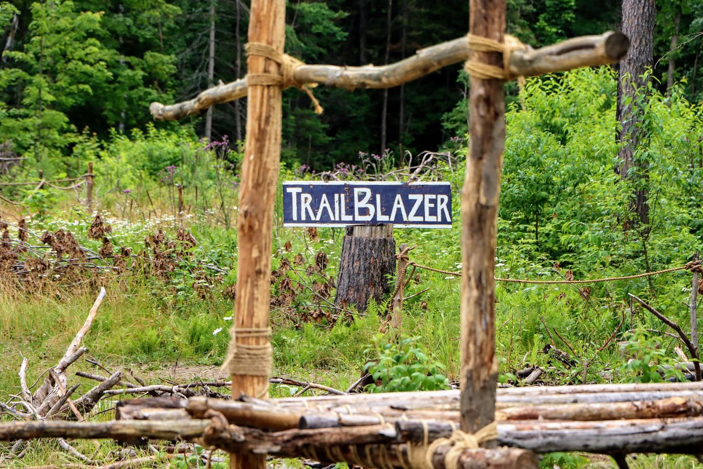 A sign that reads "TrailBlazer" hangs from a rope, suspended between two trees.