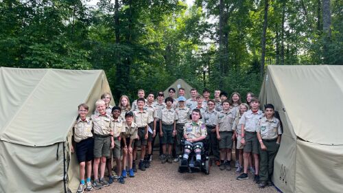 Scouts smile at the camera while standing in front of tents at a campsite