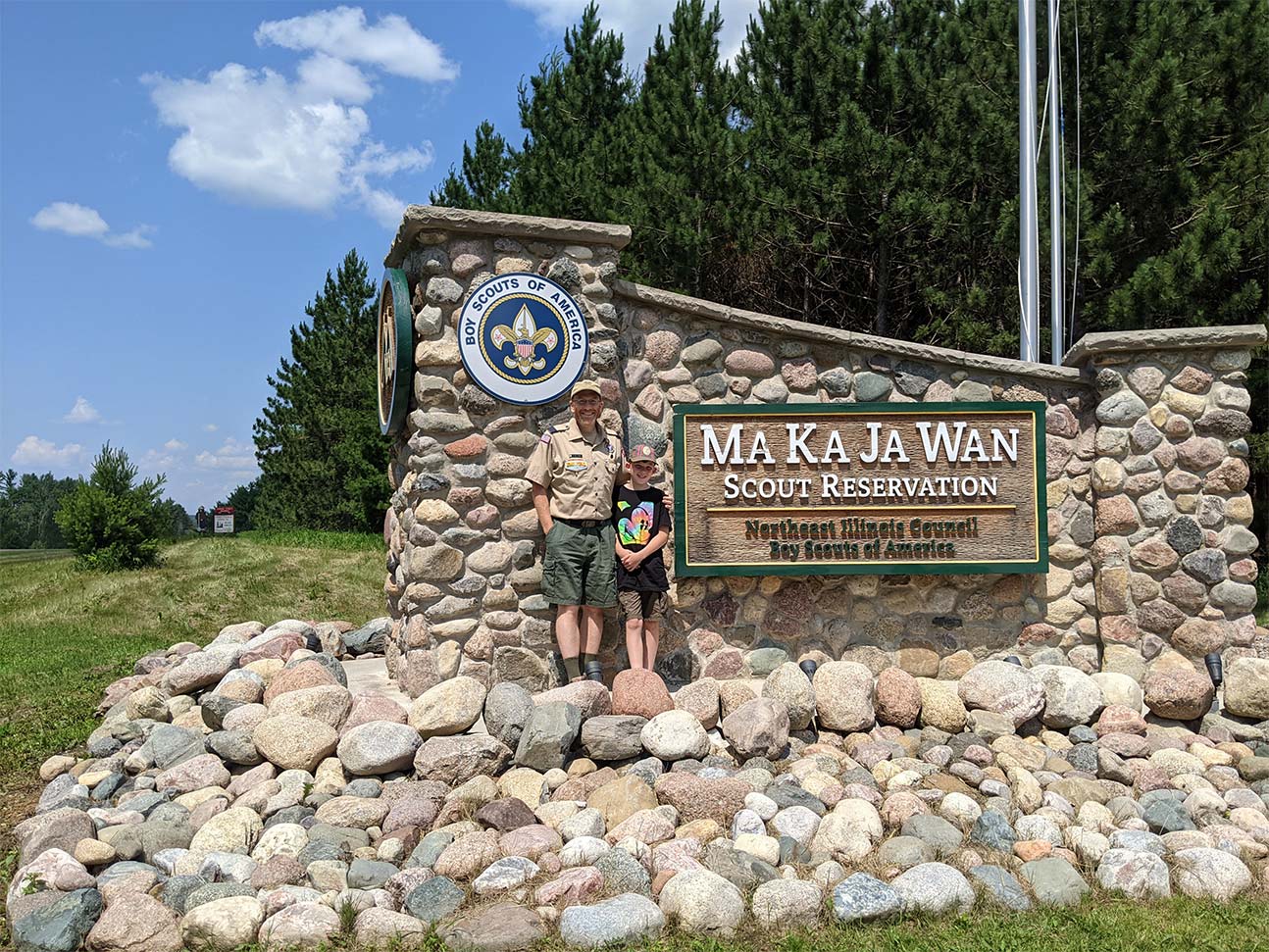 A man and a boy stand by the iconic stone welcome sign to Camp MaKaJaWan, smiling.