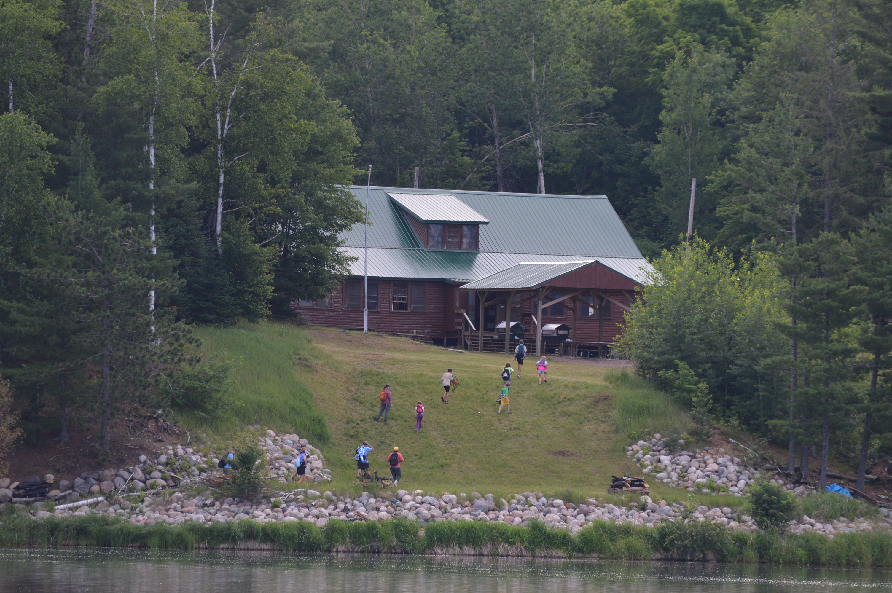 The dining hall as seen from the lake, a large log structure with deep green roof enclosed on all sides by tall trees.