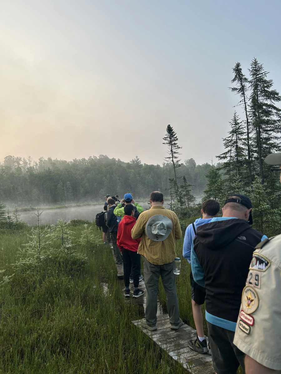 People walk in a line near the shore of the bog, with misty forest visible across the bog in the distance
