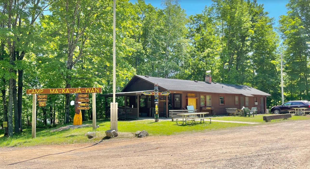 The Ma-Ka-Ja-Wan Welcome Center is a modest log cabin below a canopy of green trees
