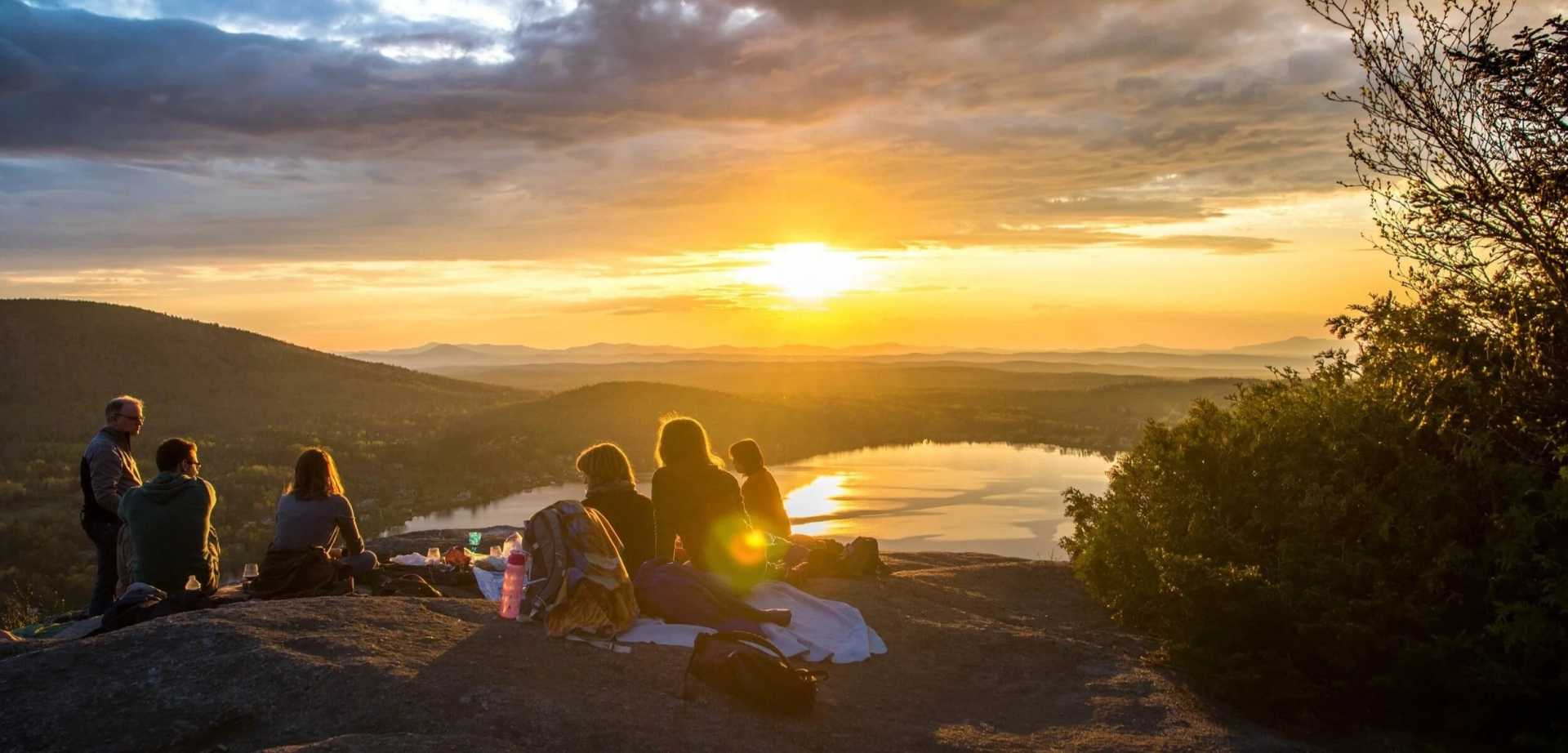 A group of campers sit atop a plateau with the sun setting behind their camp site