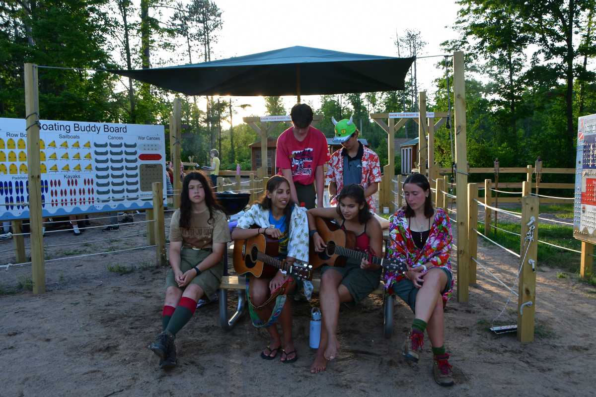 A group of girls and boys sit on a bench at camp, playing instruments and singing