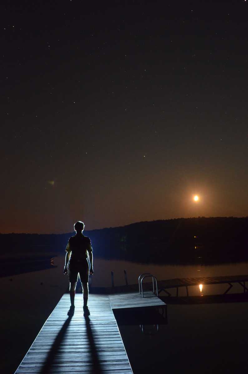 A boy scout stands on a pier at night, looking out over the water lit up by the moonlight.