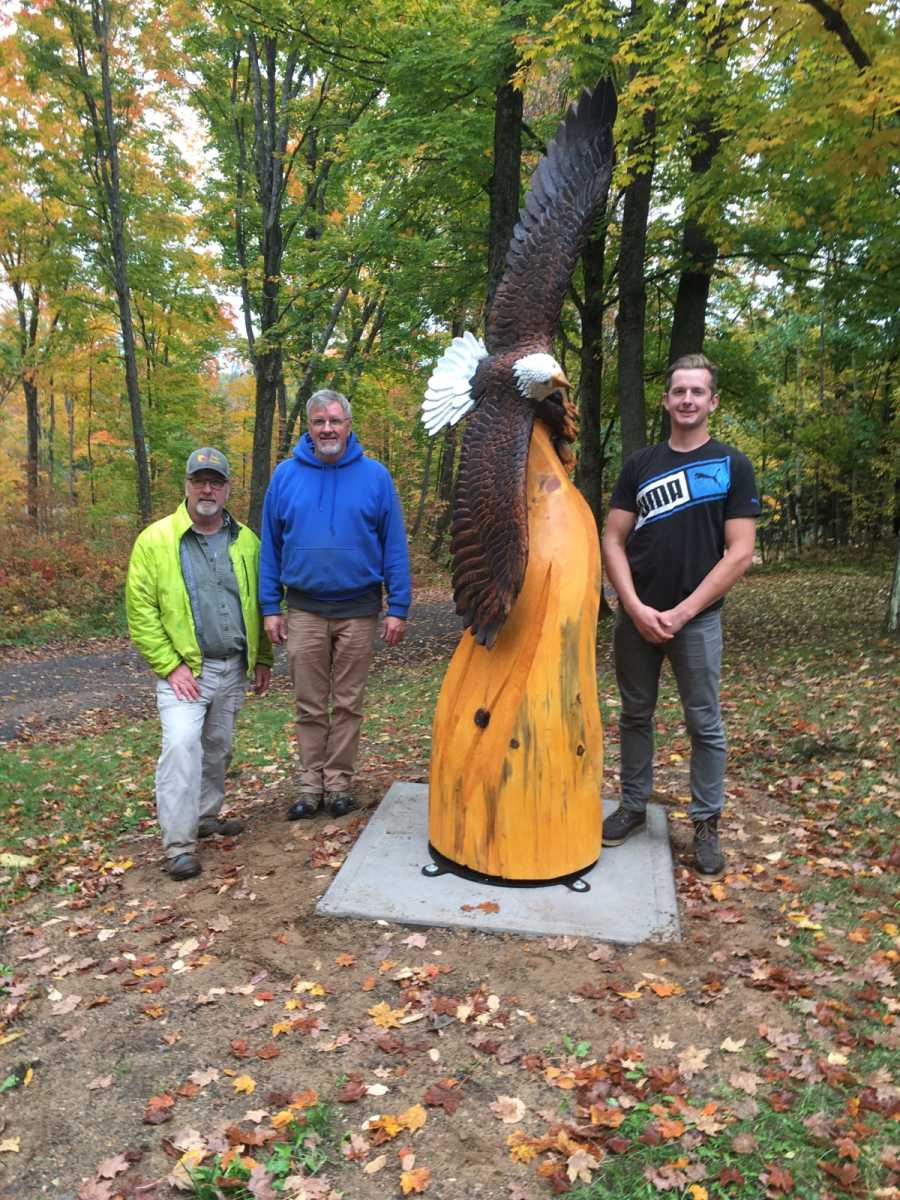 Three adults stand around an 8 foot tall wooden statue depicting a bald eagle soaring