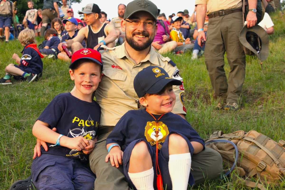 An adult scout leader holds his sons in his lap while they sit in the grass during a Family Adventure Camp event