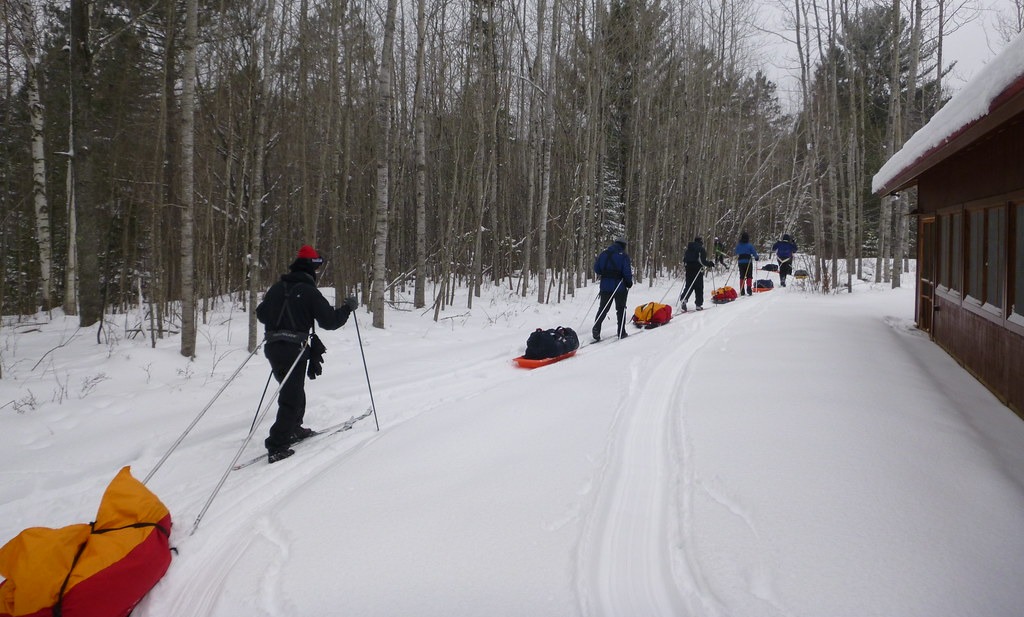 A line of people skii through thick snow with a forest visible behind them.